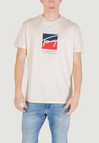 Beżowy bawełniany T-shirt z kwadratowym logo w kolorach czerwonym, białym i niebieskim, z krótkimi rękawami i luźnym krojem. Napis "Tommy."