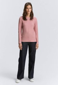 Jasnoróżowa bluzka z długim rękawem o prążkowanej teksturze, zestawiona z ciemnymi szerokimi spodniami jeansowymi i białymi trampkami, stojąca na neutralnym tle.