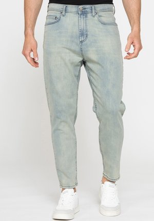 Uomo che indossa jeans azzurri a gamba stretta e sneakers bianche in piedi davanti a uno sfondo semplice.