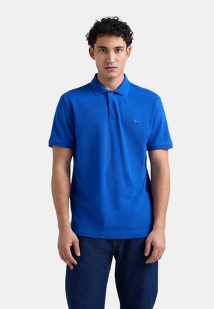 Giovane uomo con capelli ricci che indossa una polo a maniche corte di un blu vivace e jeans blu scuro, in piedi contro uno sfondo bianco.