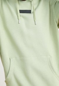 Felpa con cappuccio verde chiaro realizzata in tessuto morbido, con tasca frontale e etichetta nera con il logo. Include coulisse regolabili con punte in metallo.