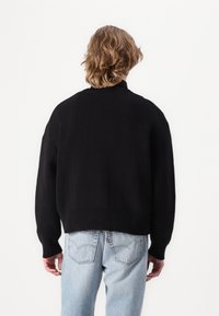 Suéter preto de malha com um colarinho alto canelado e ombros largos e caídos. Tecido texturizado e um corte descontraído, combinado com jeans azul claro.