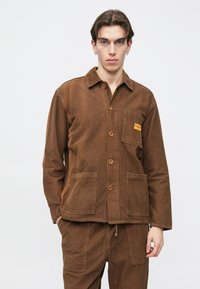 Brun kordfløyelsjakke med knappelukking foran, med to lommer foran og et lite logopatch på venstre bryst. Ensfarget krage og synlig tekstur.