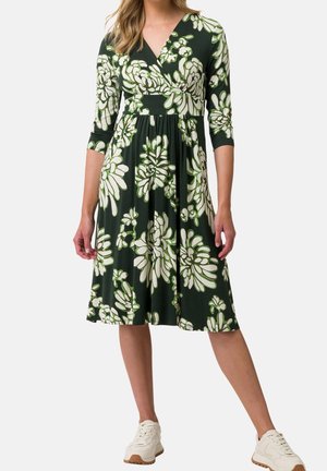 Robe portefeuille fleurie en vert foncé, ornée de grandes fleurs blanches et vert clair, manches trois-quarts, taille cintrée et jupe fluide.