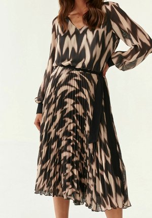 Frau trägt plissiertes Kleid mit V-Ausschnitt und langen Ärmeln, abstraktem schwarz-beigem Zickzackmuster und schwarzem Taillenband auf hellem Hintergrund.