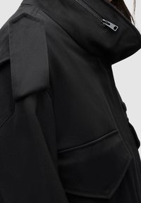 Zwarte jas met een hoge kraag, ritssluiting, opgestikte zakken en schouderepauletten, gemaakt van een getextureerde stof.