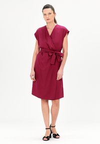 Vestido de envoltura burdeos con mangas cortas, cinturilla ajustable y longitud por debajo de la rodilla. Tejido suave con un brillo sutil. Tiras de tobillo negras.