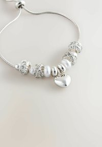 Srebrna bransoletka z zawieszkami, z białą perłą i kryształowymi koralikami, z sercową zawieszką i elastycznym, teksturowanym łańcuszkiem.