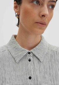 Camicia bianca con un motivo a griglia nera, dotata di bottoni neri e un tessuto testurizzato. L'orecchio del modello è decorato con un orecchino d'argento.