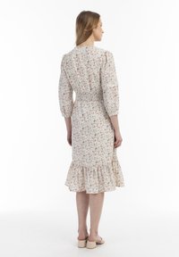 Blumenmusterkleid mit dreiviertel Ärmeln, gerafftem Taillenbund und Rüschensaum in hellem Beige und subtilen bunten Mustern. Dazu passende Sandalen.