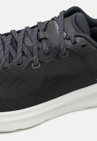 Schwarzer Sportsschuh mit einem strukturierten Stoff-Obermaterial, grauen Schnürsenkeln und einer weißen gemusterten Sohle. Auffällige Markenkennzeichnung an der Ferse und an der Seite.