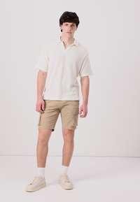 Kortermet, kremfarget polo-skjorte i strukturert stoff, kombinert med beige cargo-shorts med flere lommer og hvite sneakers.