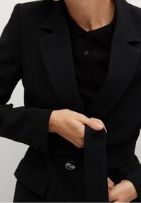 Blazer negro ajustado con hombros estructurados, solapas de muesca y una textura suave. Presenta un cierre de botón y un detalle de cinturón atado.