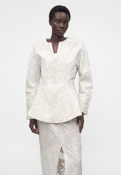 Γυναίκα που φοράει λευκό, υφασμάτινο μακρυμάνικο σακάκι με ζώνη πεπλούμ και αντιστοιχία σε φούστα μήκους μέχρι το γόνατο με εμπρόσθια σχισμή.