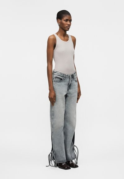 Γυναίκα που φοράει αμάνικο μπεζ ριμπ τοπ και γκρι ψηλόμεσο τζιν με φαρδιά πόδια και πλαινά κορδόνια, στέκεται μπροστά σε λευκό φόντο.