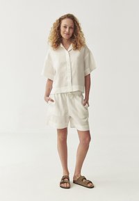 Wit linnen shirt met korte mouwen, knoopsluiting, gecombineerd met bijpassende shorts met een elastische taille en zijzakken. Bruine sandalen.