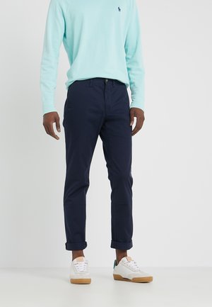 Homme portant un pantalon bleu marine retroussé, une chemise à manches longues bleu clair et des baskets blanches avec des semelles en caoutchouc, debout sur un sol blanc.