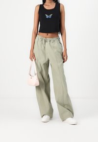 Mujer vistiendo un top corto negro con estampado de mariposas, pantalones sueltos de color verde claro, zapatillas blancas, sosteniendo un pequeño bolso de mano rosa claro frente a un fondo blanco.