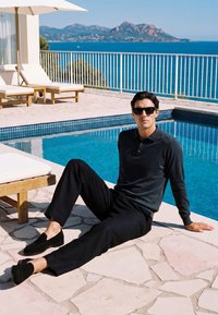 Homme en tenue noire et lunettes de soleil assis au bord d'une piscine en pierre avec vue sur l'océan et la montagne en arrière-plan par une journée ensoleillée.