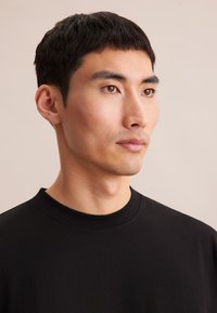 T-shirt di cotone nero con girocollo, dotato di maniche corte e una texture liscia. Indossato da un modello con capelli corti scuri, su uno sfondo chiaro.