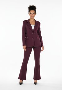 Dopasowany bordowy garnitur z jednorzędową marynarką, dwoma przednimi kieszeniami i rozszerzanymi spodniami, w połączeniu z białym topem i czarnymi butami.