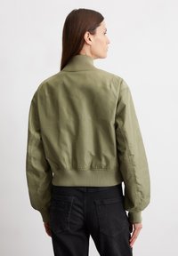 Olivgrüne Bomberjacke mit geripptem Kragen und Bündchen, die eine glatte, leicht glänzende Textur und eine taillierte Passform aufweist. Rückansicht dargestellt.