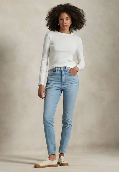 Vrouw met krullend haar die een wit shirt met lange mouwen, lichtblauwe spijkerbroek en witte sneakers draagt, staand voor een effen achtergrond.