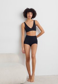 Schwarzes BH- und hochgeschnittenes Slip-Set aus dehnbarem Material. Der BH hat einen tiefen V-Ausschnitt. Das Model steht barfuß neben einem geschwungenen, hellen Sofa.