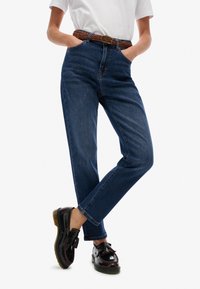 Jeans de denim de alta cintura en azul oscuro con pierna recta, que incluyen un cinturón trenzado marrón y zapatos negros con detalles de borlas.