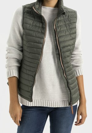 Personne portant un gilet matelassé vert sur un pull en tricot gris clair avec les manches retroussées et un jean bleu, debout devant un fond uni.