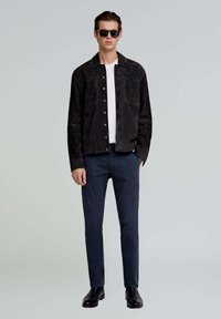 Zwarte suède jas met zilverkleurige hardware, gedragen over een wit t-shirt, gecombineerd met donkere broeken en zwarte leren laarzen. Model draagt een zonnebril.