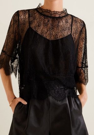 Schwarzes Spitzen-Top mit kurzen Ärmeln und Blumenmuster, getragen über einem schwarzen Camisole, kombiniert mit hoch taillierter schwarzer Lederhose, Hände in den Taschen.