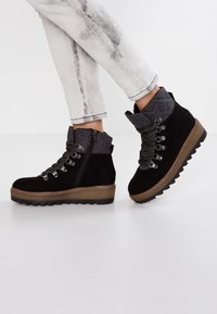 Stivali ankle in suede neri con dettagli grigio trapuntati, suola in gomma testurizzata, ganci in metallo e zip. Sono indossati jeans bianchi con effetto usurato.