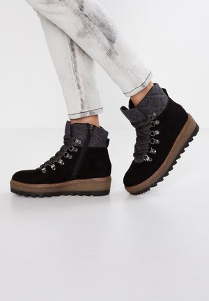 Botas de tobillo de ante negro con acentos acolchados en gris, suela de goma texturizada, ganchos de metal y detalle de cremallera. Se llevan jeans blancos desgastados.