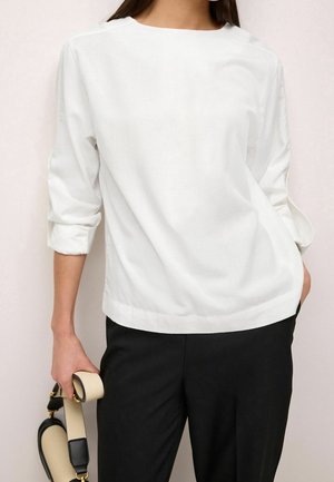 Person wearing a white long-sleeve shirt with rolled cuffs and black pants, holding a beige and black handbag by the strap.