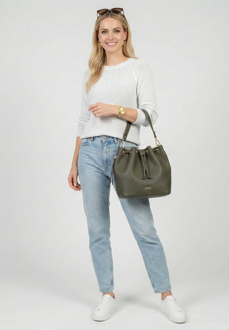 Lachende vrouw in een witte trui en lichte spijkerbroek houdt een olijfgroene leren buideltas met schouderriem vast, draagt witte sneakers en een zonnebril.