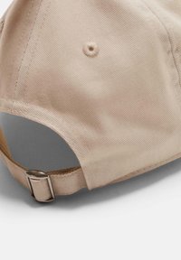 Casquette en coton beige avec une visière incurvée, tissu texturé et une sangle métallique ajustable à l'arrière. Détail de couture renforcé visible.