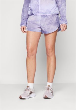 Personne portant un short tie-dye violet clair et une veste assortie, des chaussettes blanches et des chaussures de sport rose clair, debout sur un sol uni.