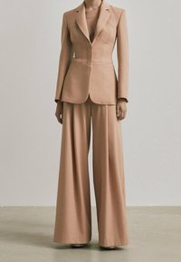 Completo su misura di un leggero beige, caratterizzato da una giacca aderente con cuciture strutturate e pantaloni a gamba larga, entrambi realizzati in un tessuto liscio.