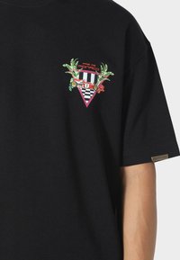 Mustat puuvillat-paita, jossa on värikäs graafinen kuvio rinnassa, jossa on palmunlehtiä, ruutukuosi ja vintage-auton muotoilu.