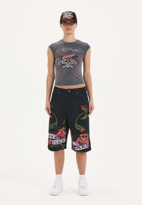 Camiseta gris corta con gráfico de calavera, combinada con shorts de mezclilla negros que presentan diseños florales coloridos y texto. Zapatillas blancas y gorra negra.