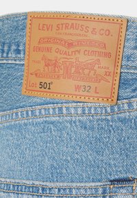 Farkut denim, joissa on ruskea nahkapaikka, jossa on punaiset ja mustat tekstit, hahmotelmat hevosista ja ompeleet reunoilla.