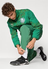 Zelena trenirka s kamuflažnim vzorcem, hlače z zadrgo in Puma logo. Model prilagaja rob hlač medtem ko nosi črne športne čevlje in bele nogavice.
