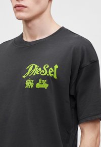 Νέος άνθρωπος φοράει μαύρο μπλουζάκι Diesel με φωτεινό πράσινο κείμενο "Diesel since 1978 fine quality" στο στήθος.