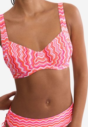 Frau trägt Bikini mit rosa, orange- und weißem Zickzack-Muster, eine Hand ruht auf der Hüfte.