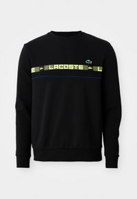 Svart langermet sweatshirt med gul grafisk tekst og grønn krokodillelogo over brystet, med blå pipingdetalj under.