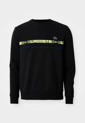 Sweat-shirt noir à manches longues avec un texte graphique jaune et un logo de crocodile vert sur la poitrine, présentant un détail de piping bleu en dessous.