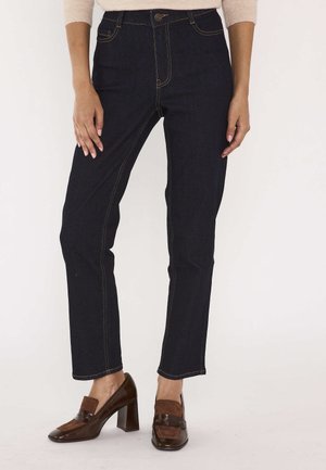 Femme portant un jean droit bleu foncé et des mocassins marron à talons, debout devant un fond uni.