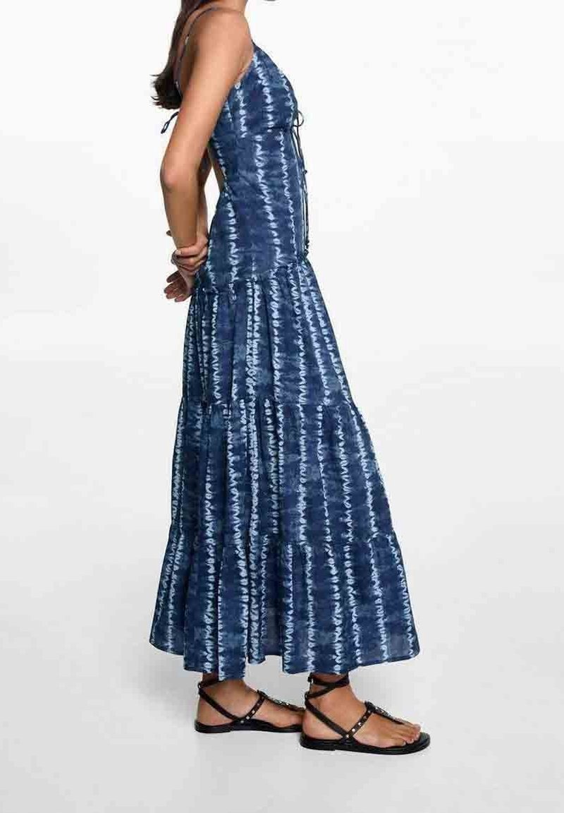 Femme debout portant une robe maxi sans manches à effet tie-dye bleu avec une jupe à étages et des sandales plates noires à brides à la cheville.