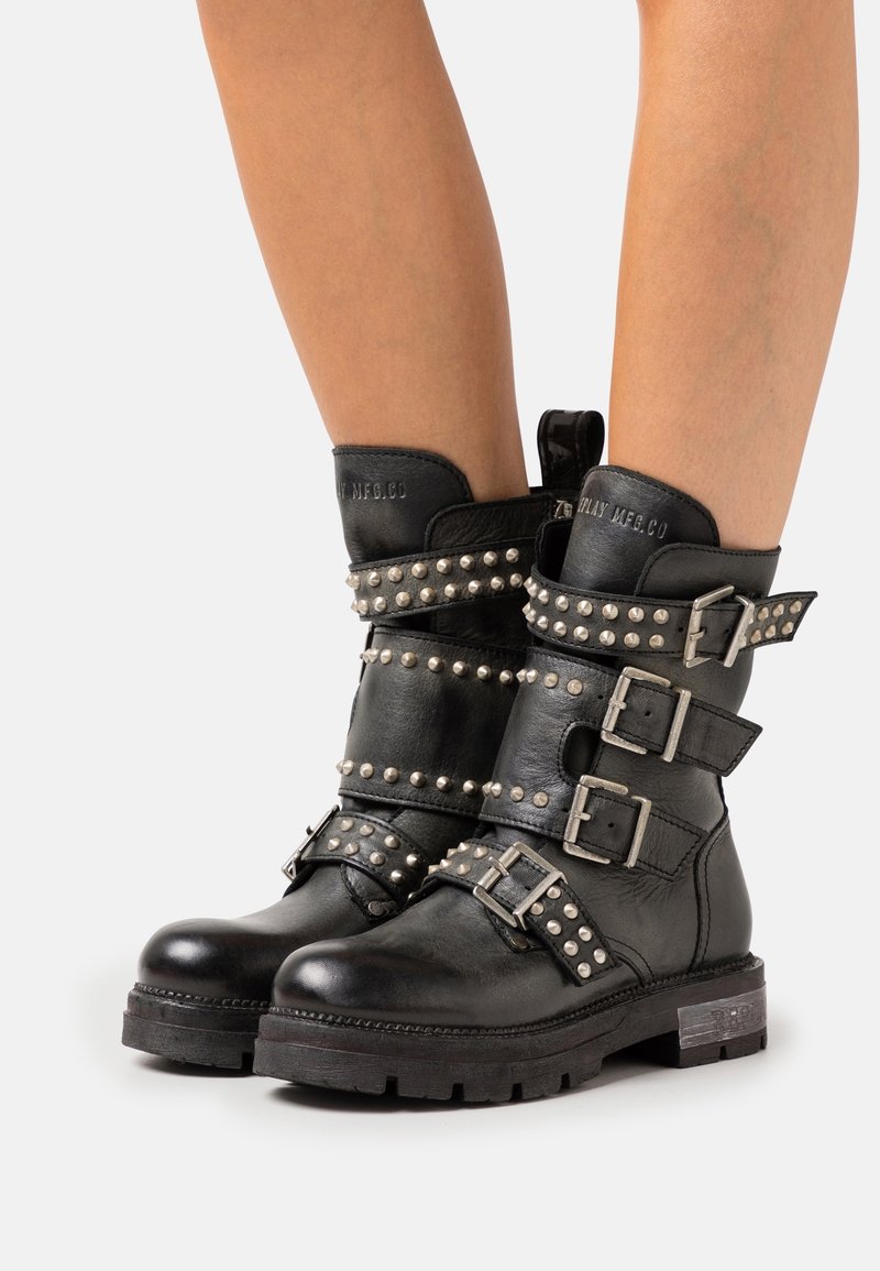 Botas de tobillo de cuero negro con acentos de tachuelas plateadas, múltiples correas ajustables y una suela gruesa. Logotipo exhibido en el talón.